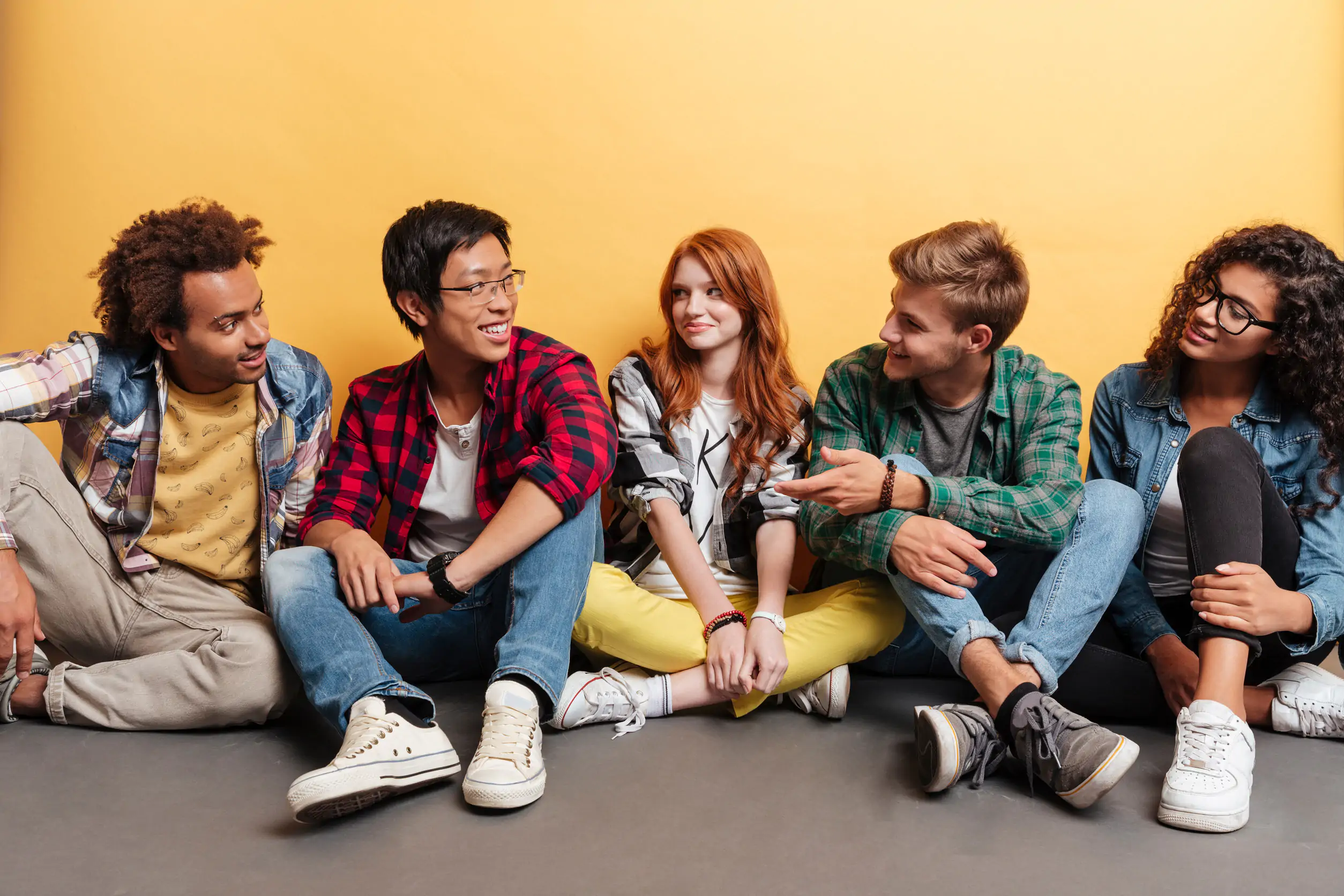 Multiethnic group of smiling young people sitting on the floor and talking over yellow background